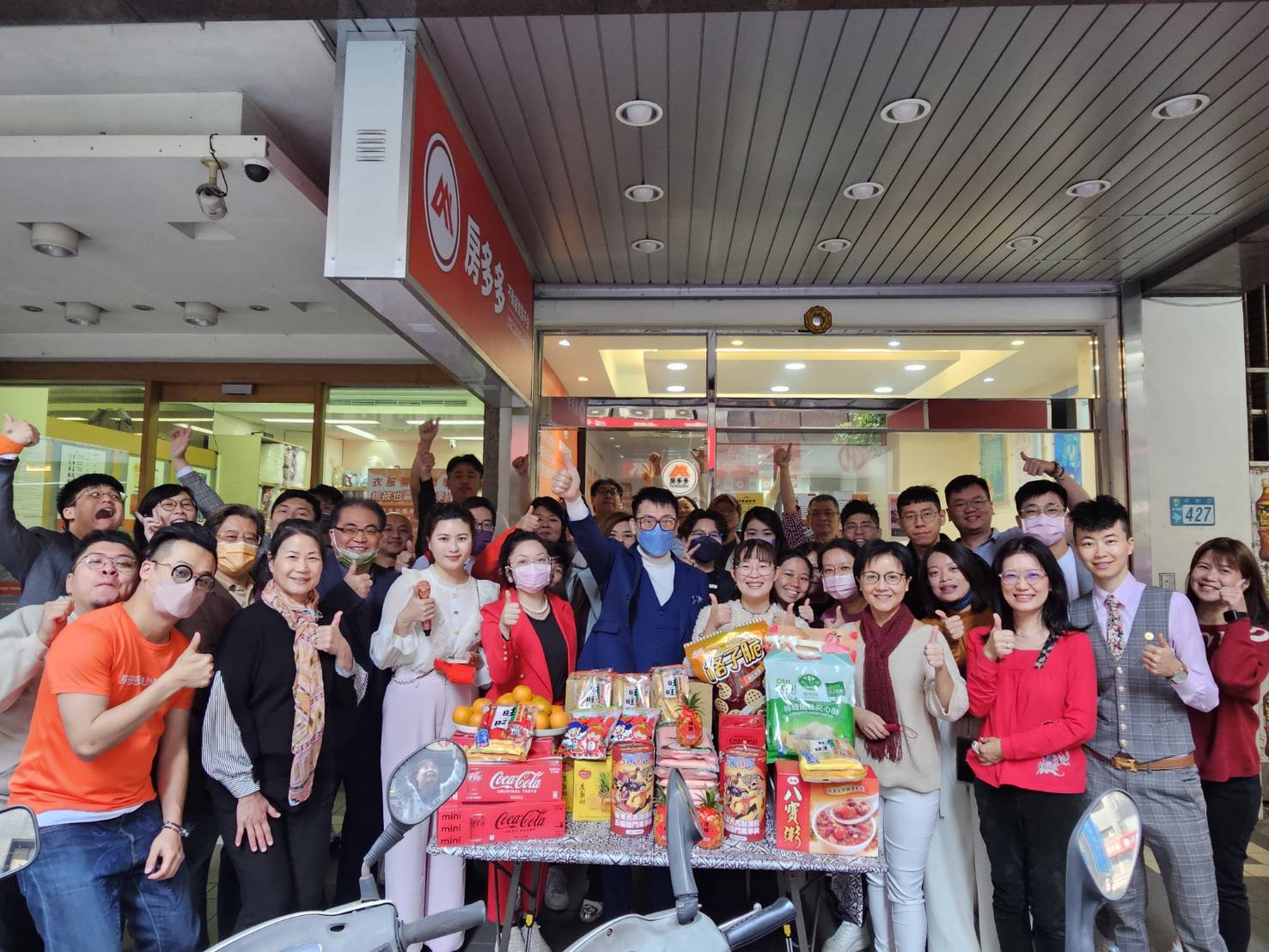一群房多多團隊和客戶在店面前圍繞擺滿零食與飲料的桌子慶祝活動，展現房屋投資社群的熱情