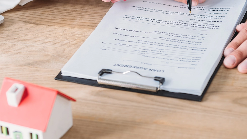 桌面上擺放迷你房屋模型和貸款合約文件，有人拿筆閱讀，象徵了解買房注意事項的過程