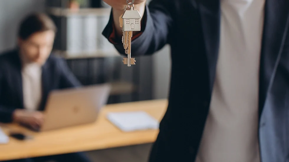 一位身穿西裝的人手中拿著帶有小房子吊飾的鑰匙，背後有人在辦公桌前工作，象徵房地產投資課程中的實務交易