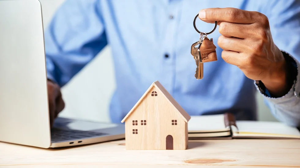 一名男士坐在筆電前拿著房屋鑰匙，桌上有木製小房子模型，呈現投資房地產與購屋的概念