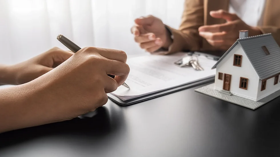 兩人在桌前討論並簽署文件，旁邊放著縮小的房屋模型，象徵投資房子或房地產交易