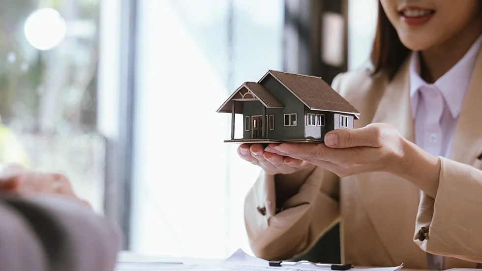 一位女性雙手捧著小型房屋模型交給對方，象徵投資房屋的討論或諮詢